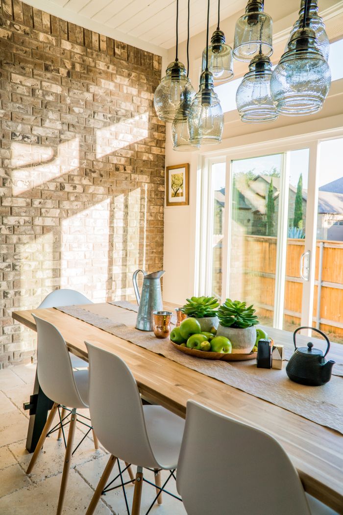 ours-journey Sunlit dining room with contemporary decor and a rustic brick wall, featuring stylish lighting and a wooden table.