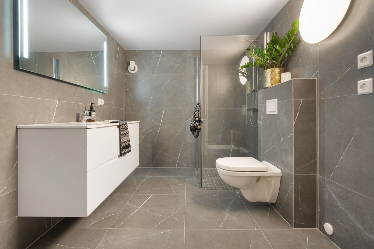 gallery-2 Stylish gray tiled bathroom featuring a shower, toilet, and illuminated mirror.